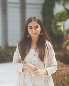 a woman with long brown hair wearing a white dress and earrings standing in front of a house