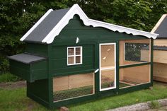 a green chicken coop with two windows and a white roof on the grass in front of some trees