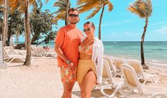 a man and woman standing on the beach in front of palm trees with chairs around them