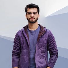 a man with glasses standing in front of a blue and white background wearing a purple jacket