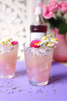 two glasses filled with ice and flowers on top of a purple table next to a bottle