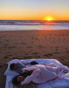 a woman laying on top of a bed under a blanket next to the ocean at sunset