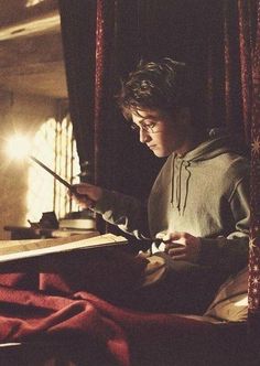 a young man sitting on top of a bed next to a table holding a book