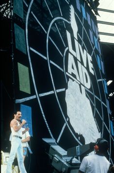 two men on stage with microphones in front of a large screen that says live aid