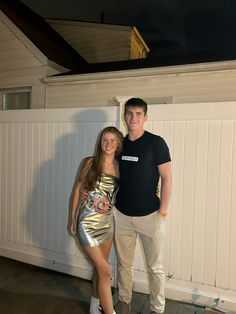 a man and woman standing next to each other in front of a white fence at night