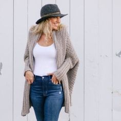 a woman wearing jeans and a hat is standing in front of a white wall, talking on her cell phone