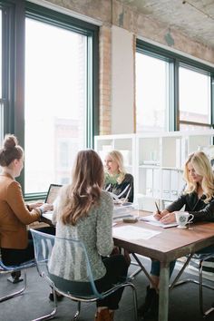 four women sitting at a table with laptops and papers in front of large windows