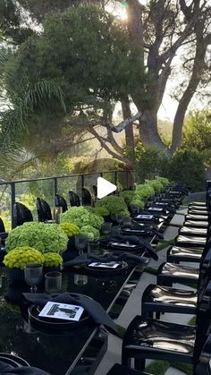 a long table with many black chairs and plants on it in front of some trees