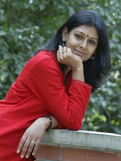 a woman in a red dress leaning on a brick wall with her hand under her chin