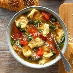 a bowl of pasta and spinach soup on a cutting board next to two croissants