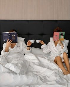 two women in white robes are reading books and drinking coffee while laying on a bed