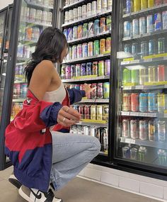 a woman squatting down in front of a refrigerator full of sodas and drinks