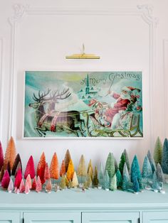 there are many different colored christmas trees on the shelf in front of the painting and other decorations