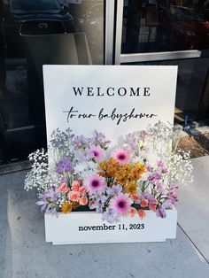 a welcome sign sitting on the side of a building with flowers growing out of it