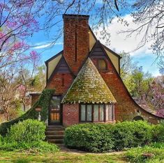 a brick house with a thatched roof surrounded by green bushes and pink flowering trees