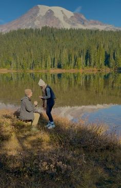 two people sitting on a bench near a lake with a mountain in the back ground