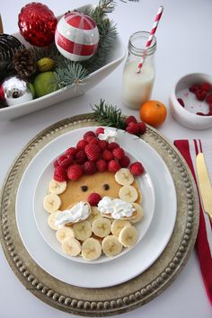 a white plate topped with pancakes covered in banana slices and raspberries next to a bowl of fruit