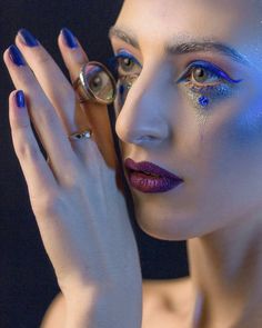 a woman with blue and silver makeup holding her hand up to her face