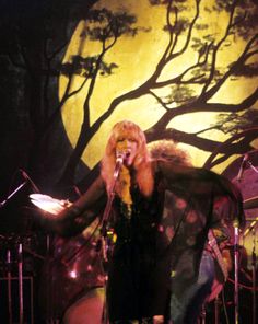 a woman standing on stage in front of a tree with her hands out to the side