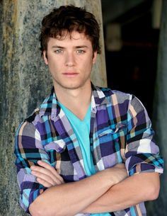 a young man with his arms crossed standing in front of a tree and looking at the camera