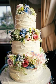 a three tiered wedding cake with flowers on the top and bottom, sitting on a table in front of a window