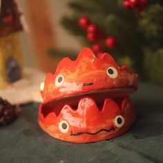 a red ceramic object with eyes and mouth sitting on a table next to a christmas tree