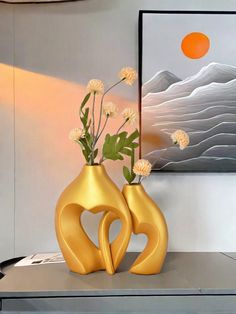 two gold vases with flowers in them on a table next to a painting and lamp