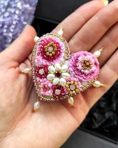 a hand holding a pink heart shaped brooch with pearls and flowers on the side