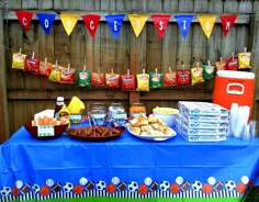 a blue table topped with lots of food next to a wooden fence covered in flags