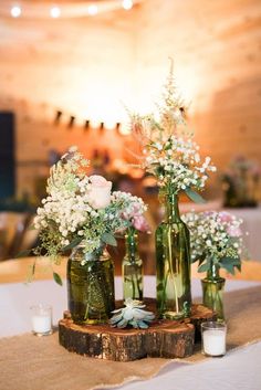 three green vases with flowers are sitting on a wooden slab in front of a white table cloth