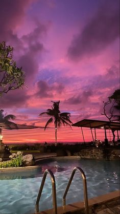 an outdoor swimming pool at sunset with palm trees and umbrellas in the foreground