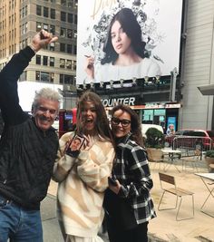 two women and a man pose for a photo in front of a large advertisement on the side of a building