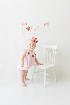 a baby girl standing in front of a white chair with a name banner above her head