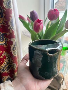 a hand holding a green vase with flowers in it next to a red and white curtain