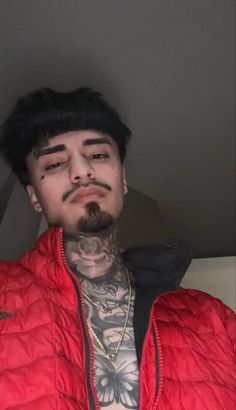 a man with black hair and tattoos wearing a red puffy jacket looking at the camera