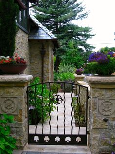 an iron gate with flowers and potted plants at the entrance to a stone house