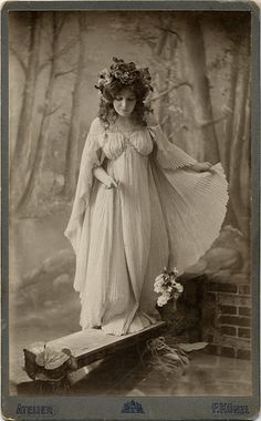 an old fashion photo of a woman with flowers on her head and dress, standing on a log in the woods