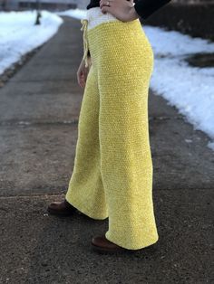 a woman standing on a sidewalk wearing yellow knitted pants and brown shoes with her hands in her pockets