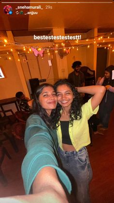 two people taking a selfie in a room with string lights on the ceiling and chairs