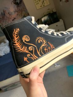 someone is holding up their black and orange painted shoe with fern designs on the side