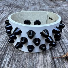 White leather studded punk bracelet with 3 rows of little cone studs. Studs measure 3/8" across and around 3/8" tall. Studs made with black enameled steel. Snaps made from matte black brass. Please note the the snaps have a nickel base layer. Cuff is 1.5" wide. Made with 7-8 oz (1/8", 3mm) white leather. Punk Black Bracelet With Silver Studs, Black Edgy Wristband With Studs, Black Leather Punk Bracelet With Silver Studs, Punk Leather Bracelet With Studs, Edgy Leather Bracelets With Studs, Punk Style Studded Leather Bracelet, Black Rocker Bracelets With Rivets, Black Rocker Bracelet With Studs, Black Leather Bracelet With Studs For Rock Style