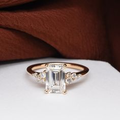 an emerald cut diamond ring on top of a white surface with brown fabric in the background