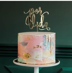 a multi - colored cake with gold lettering on top sits on a white pedestal in front of a green wall