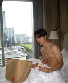 a shirtless man sitting on a bed with a bag and drink in his hand