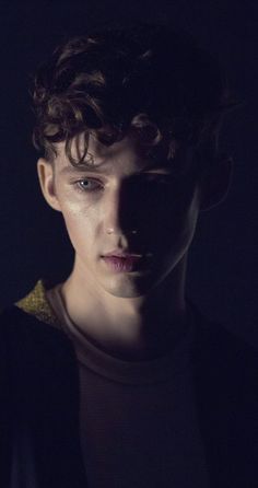 a man with curly hair standing in the dark wearing a black jacket and gold trim