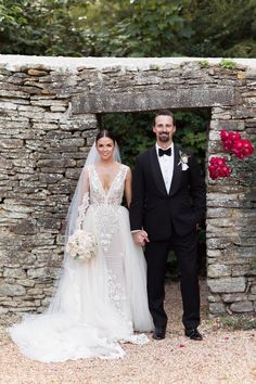 a bride and groom standing in front of a stone wall with red flowers on it