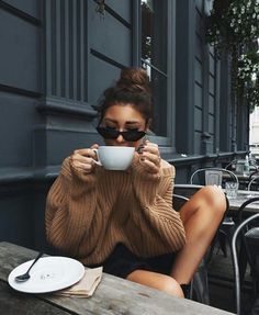 a woman sitting at an outdoor table drinking from a coffee cup with her eyes closed