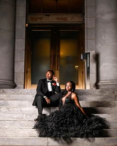 a man and woman sitting on the steps in front of a building wearing tuxedos