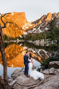 a bride and groom standing on the rocks near a mountain lake with their reflection in the water