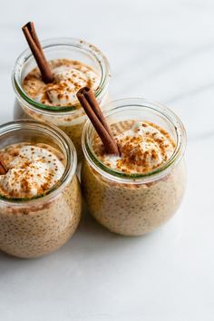 three small jars filled with food and topped with cinnamon stick garnishments on a white surface